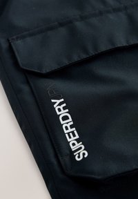 Tessuto nero con una texture liscia presenta una tasca frontale. Logo bianco "SUPERDRY" stampato verticalmente sul lato.