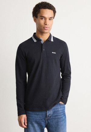 Svart langermet polo skjorte med en kontrasterende stripekrage. Har et lite hvitt logo på brystet og en avslappet passform. Kombinert med blå denimjeans.