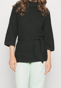 Suéter negro de punto con cuello alto, mangas 3/4 y un cinturón de tela en la cintura. El acabado texturizado añade profundidad. Combinado con pantalones verde claro.