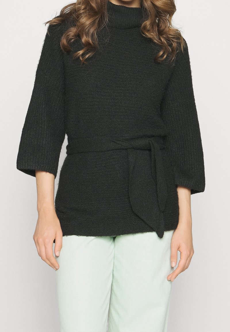 Suéter negro de punto con cuello alto, mangas 3/4 y un cinturón de tela en la cintura. El acabado texturizado añade profundidad. Combinado con pantalones verde claro.