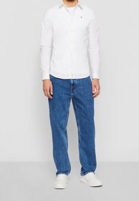 Uomo che indossa una camicia bianca a maniche lunghe con bottoni, jeans blu dritti e sneaker bianche, in piedi contro uno sfondo semplice.