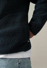 Pullover in pile di un blu navy scuro con una superficie strutturata e tasche laterali, caratterizzato da un design aderente e indossato sopra una T-shirt bianca.