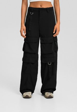 Pantalones cargo negros de pierna ancha con múltiples bolsillos con solapa y detalles de anillo en D, usados con zapatillas beige, top blanco corto visible en la cintura.