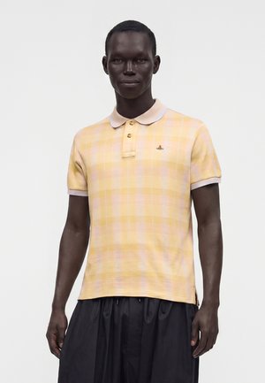 Homme portant un polo à manches courtes à carreaux jaune et rose avec un petit logo sur la poitrine et un pantalon noir ample, debout devant un fond uni.