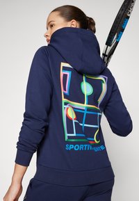 Námornícky modrá mikina vyrobená z bavlnenej zmesi, s farebnou geometrickou grafikou na chrbte a nápisom "SPORTIVAMENTE." Hladká textúra, priliehajúce manžety.