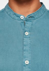 Chemise bleu clair avec col montant, fermeture boutonnée à l'avant et texture subtile. Comprend un seul bouton au col et des surpiqûres visibles.