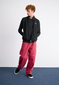 Melna flīsa zīmu vestē virs melnas garām piedurknēm krekla, apvienota ar brīvām sarkanām biksēm un melniem slip-on apaviem uz zila paklāja.