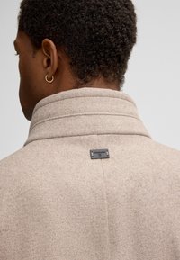 Close-up van een persoon met kort, krullend haar die een beige wollen jas draagt met een klein zwart rechthoekig label op de kraag aan de achterkant en een gouden ringoorbel.