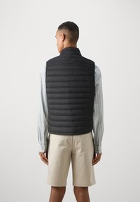 Vesta puf neagră cu model matlasat orizontal, guler înalt și fără mâneci, purtată peste o cămașă cu dungi albastre deschise și pantaloni scurți bej.