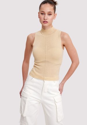 Jeune femme aux cheveux attachés en chignon, portant un haut beige sans manches à col roulé et un pantalon cargo blanc, debout les mains dans les poches.