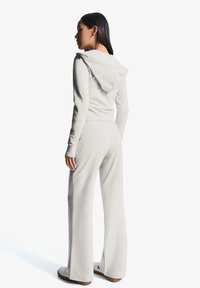 Eine junge Frau steht mit dem Rücken zum Betrachter, trägt einen hellgrauen Kapuzenpulli und eine passende Hose, vor einem minimalistischen weißen Hintergrund.
