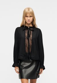 Γυναίκα με μακριά ξανθά μαλλιά που φοράει μαύρη μπλούζα με δαντέλα και βελούδινη γραβάτα, καθώς και μαύρη δερμάτινη μίνι φούστα, μπροστά σε απλό φόντο.