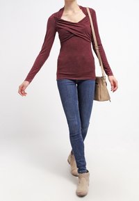 Långärmad vinröd topp med draperad halsringning och samlade detaljer, tillsammans med blå tajta jeans och beige ankelstövlar, bärande på en tan axelväska.