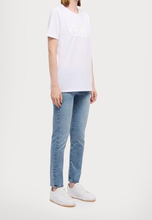 Personne portant un t-shirt blanc à manches courtes avec un logo subtil, un jean bleu clair et des baskets blanches, debout devant un fond uni.