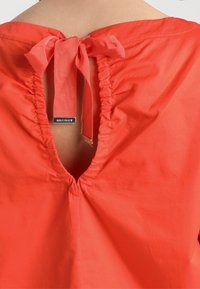 Vêtement rouge noué au niveau de l'encolure arrière avec un nœud, montrant une petite ouverture en forme de trou de serrure et une étiquette de marque en métal visible.