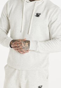 Felpa grigia chiara con tasca a marsupio, cappuccio con coulisse e logo nero. Presenta polsini a costine e un tessuto morbido e texturizzato.