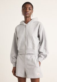 Felpa grigia corta con zip frontale e tasca a marsupio abbinata a pantaloni corti grigi. Tessuto morbido, vestibilità rilassata, dettagli di branding minimi.
