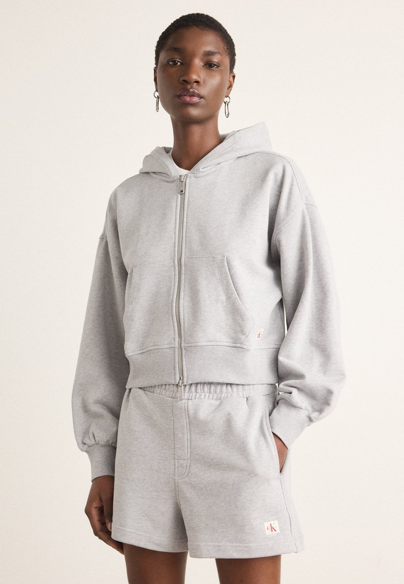 Felpa grigia corta con zip frontale e tasca a marsupio abbinata a pantaloni corti grigi. Tessuto morbido, vestibilità rilassata, dettagli di branding minimi.