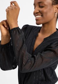 Schwarze durchsichtige Bluse aus strukturiertem Stoff, mit dezenten silbernen Streifen und Puffärmeln. Sie hat einen V-Ausschnitt und geraffte Handgelenke.
