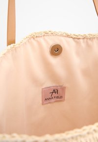 Tan tote-väska med en vävd halmtrimmning, med en mjuk persikofärgad insida och en metallknappsstängning. Inkluderar en etikett med "ANNA FIELD"-logotyp.