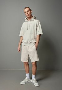 Beige bomulds hoodie med en frontlomme og snor, parret med matchende shorts. Båret med hvide sneakers og ankelsokker.