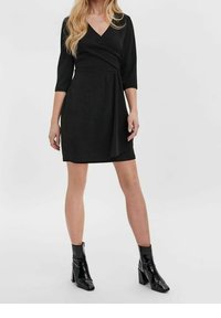 Schwarzes Wickelkleid mit Dreiviertelärmeln, einem V-Ausschnitt und fließendem Stoff, kombiniert mit glänzenden schwarzen knöchelhohen Stiefeln und Blockabsätzen.