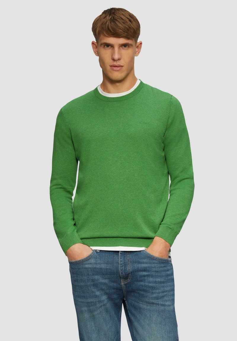 Groene gebreide trui met geribde manchetten en halslijn, gedragen over een wit T-shirt, in combinatie met blauwe denim jeans.