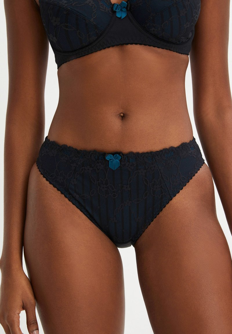 Sous-vêtement en dentelle noire ornée de motifs floraux, avec des bords festonnés et un petit nœud teal accent à l'avant. Tissu doux et texturé.