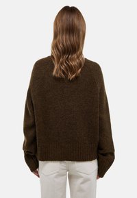 Pull oversized marron foncé avec ourlet et poignets côtelés. Fabriqué en tissu doux et texturé. Porté par-dessus un pantalon de couleur claire.