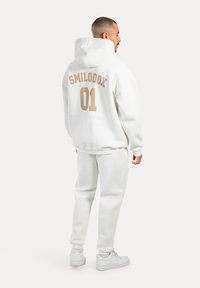 Felpa bianca con "SMILODOX 01" in beige sulla schiena, abbinata a pantaloni bianchi e sneakers bianche. Tessuto morbido, vestibilità rilassata.