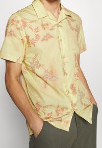 Korte mouwen shirt van lichtgele stof met een bloemenpatroon in tinten koraal en blauw, met een kraag en knoopsluiting aan de voorkant.