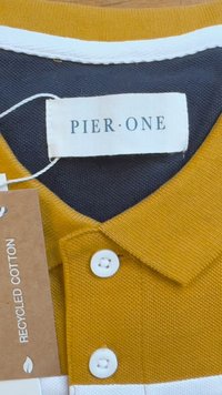 Rumena polo majica s temno modrim ovratnikom. Ima bele poudarke, tri bele gumbe in oznako, ki nakazuje, da je izdelana iz recikliranega bombaža.