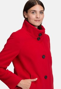 Vrouw in een felrode jas met een hoge kraag en zwarte knopen, hand in de zak, tegen een plain witte achtergrond.