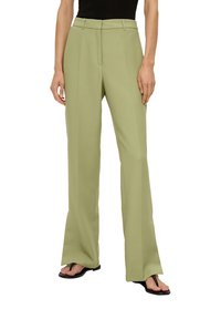 Pantalons larges vert clair en tissu doux, avec un pli sur le devant et une taille ajustée. Portés avec des tongs noires.