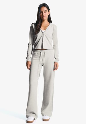 Een jonge vrouw staat zelfverzekerd in een lichtgrijze crop hoodie en bijpassende joggingbroek, gecombineerd met witte sneakers, tegen een effen achtergrond.