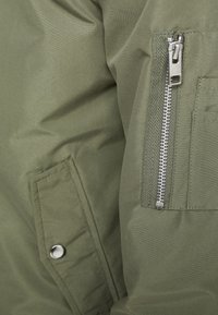Detail einer olivgrünen Jacke mit strukturiertem Stoff, einem silbernen Reißverschluss und einer Druckknopf-Tasche am linken Ärmel.