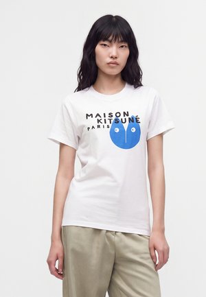 Jeune femme aux longs cheveux noirs portant un T-shirt blanc Maison Kitsuné Paris avec un motif visage de renard bleu et un pantalon beige.