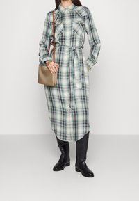 Dlouhé kostkované šaty v zelených a modrých tónech, s knoflíkovým zapínáním, dvěma náprsními kapsami, opaskem v pase a v kombinaci s černými botami do kolen.