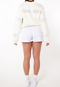 Bílý zkrácený svetr s otevřeným pletením v kombinaci se šedými šortkami. Svetr má dlouhé rukávy a kulatý výstřih.