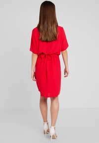 Vestido rojo hasta la rodilla con mangas sueltas, detalle de cintura atada y tejido ligero. La parte posterior presenta un acabado liso y sencillo.