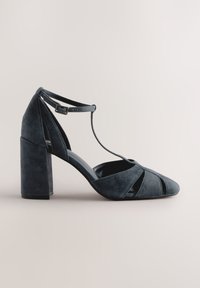 Donkerblauwe suède hoge hak met een uitgesneden ontwerp, spitse neus en enkelbandje. De hak is stevig en vierkant van vorm.