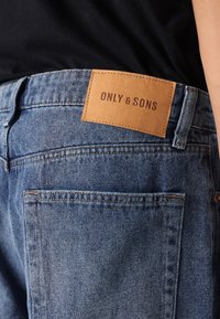 Jeansi din denim albastru cu o etichetă din piele maro pe buzunarul din spate, având cusături contrastante și un design clasic cu cinci buzunare.