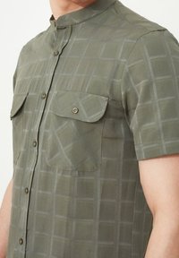 Lyhythemdikavöntöinen paita oliivinvihreänä, ruutukuviolla, kahdella läpällä varustetulla taskulla, mandariinikauluksella ja sileällä pinnalla.