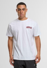 Bílá bavlněná tričko s vertikálními proužky a červeným logem "UNFAIR ATHLETICS" na levé části hrudi. Standardní design s kulatým výstřihem.