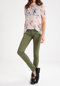 Blusa de manga corta con estampado floral, tejido transparente, en color rosa claro con acentos en negro y dorado, combinada con pantalones ajustados verde oliva y zapatos negros.