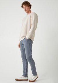 Sudadera de un beige claro con cuello redondo, combinada con unos jeans ajustados de un azul claro. Zapatillas de deporte blancas con detalles marrones completan el conjunto.