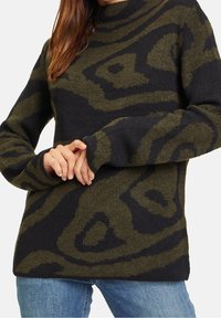 Femme portant un pull à manches longues à motifs verts foncés et noirs, les mains croisées devant, accompagné d'un jean bleu.