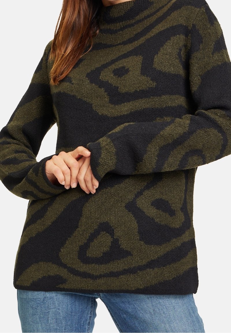 Femme portant un pull à manches longues à motifs verts foncés et noirs, les mains croisées devant, accompagné d'un jean bleu.