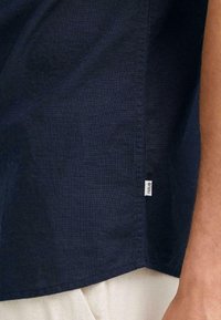 Camisa de lino azul marino con un acabado texturizado, que presenta un dobladillo recto y una pequeña etiqueta en la costura lateral.