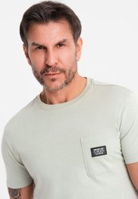 Světle zelené tričko s krátkým rukávem vyrobené z měkké látky, s náprsní kapsou a černou nálepkou s textovým detailem. Čistý, jednoduchý design.